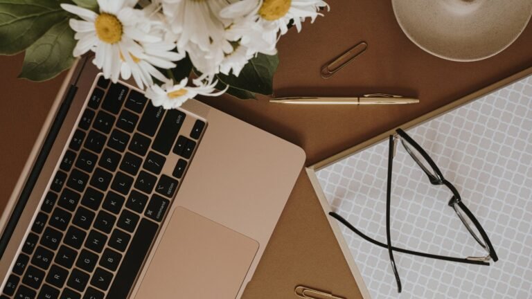 Top-down view of a brown desk with a rose-gold laptop, white daisies in a vase, a gold pen, paper clip, cup, and glasses on a patterned notebook
