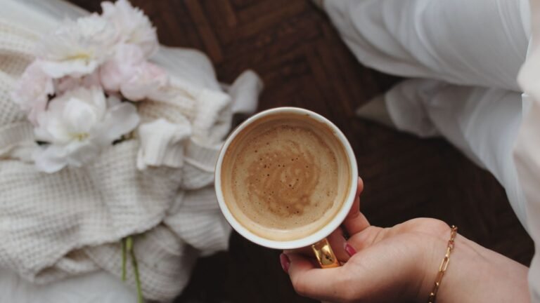 Hand holding a cup of coffee with crema, with a soft beige blanket and bouquet in the background.