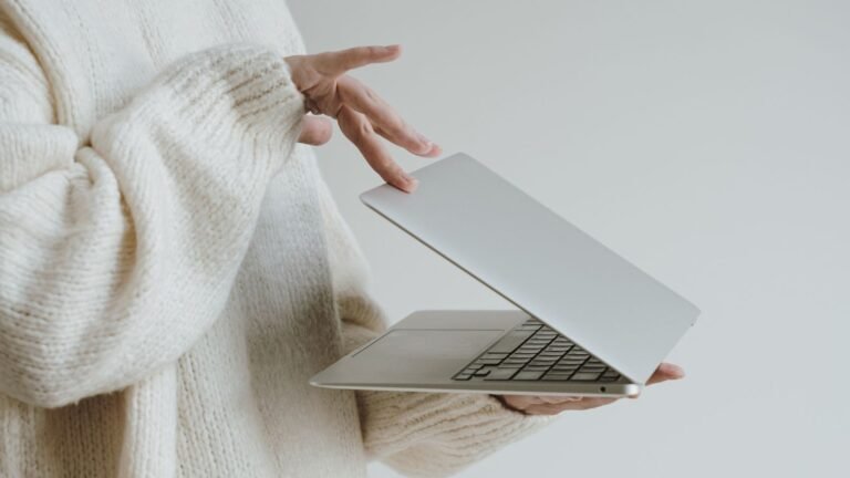 Person in a cream knit sweater lifts the lid of a silver laptop.