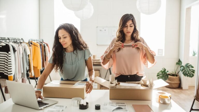 Two women packing orders at a bright clothing boutique, with racks of clothes in the background.