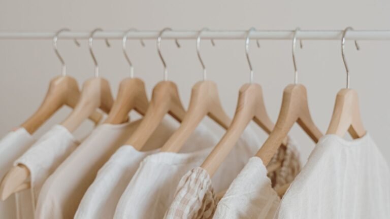 Rows of wooden hangers with light-colored shirts hanging on a metal clothes rail in a neutral closet.