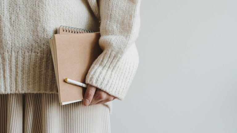 Person in a chunky cream knit sweater holds a brown spiral notebook with a white pen tucked in the cover, against a light background.