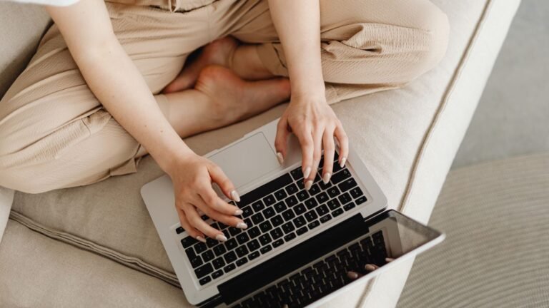 Person sitting on a beige couch typing on a silver laptop on the lap/desk surface.