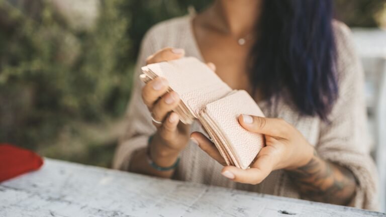 Person counts a stack of pink cash held over a weathered white table outdoors, with a beige sweater visible in the background.