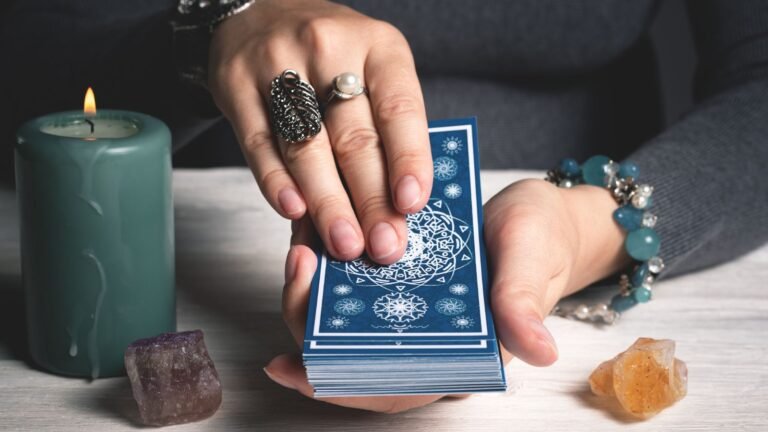 Hands holding a deck of blue-backed cards with intricate white designs, candle and crystals on a wooden table in the background.