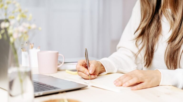 Person writing in a notebook at a tidy desk with a pink mug and a laptop nearby on a bright workspace appeneded scene.