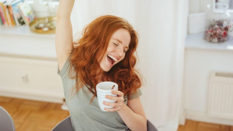 Red-haired woman wearing a light gray shirt, smiling with eyes closed, raising one arm while holding a striped mug indoors.