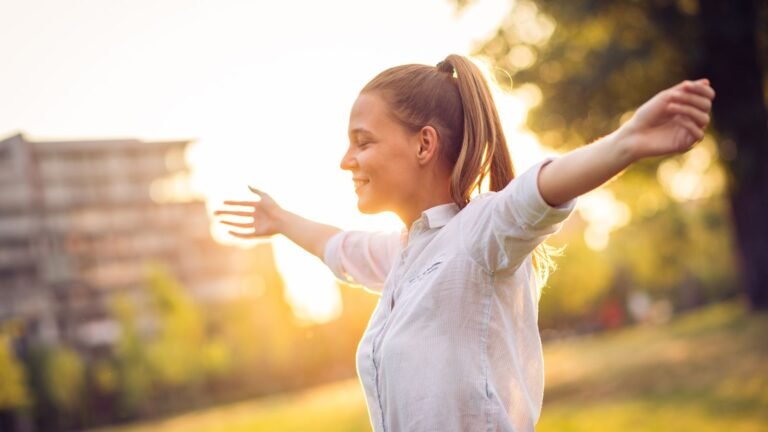 Woman standing outdoors with arms outstretched in sunset, smiling in the warm evening light.