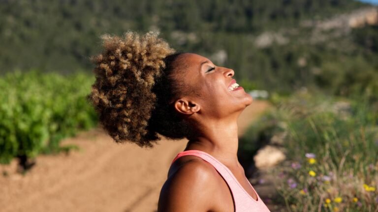 Woman with curly hair in a pink tank top tilts her head back and smiles in bright sunlight outdoors.