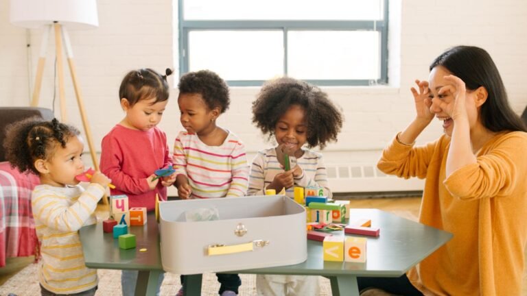 Caregiver and four young children play with colorful blocks at a table in a bright classroom.