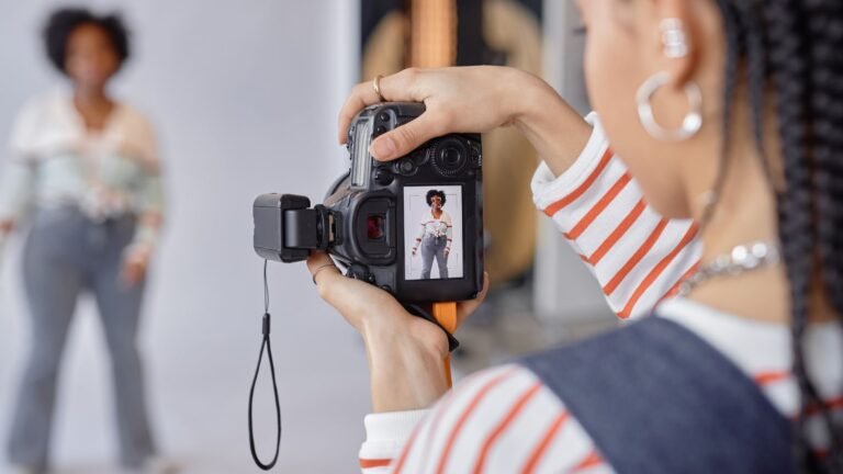 Photographer in a striped shirt shoots a model in a studio; camera LCD shows the model posing in jeans and a white top.