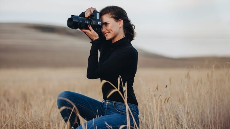 Woman in a black turtleneck and jeans crouches in a dry field, aiming a DSLR camera at eye level.
