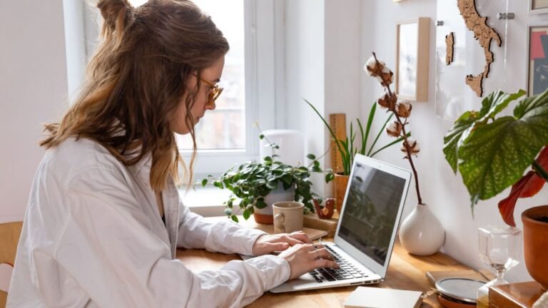 Person typing on a silver laptop at a wooden desk by a window, surrounded by green potted plants and a mug nearby.