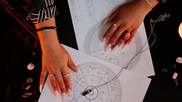 Person's hands with long manicured nails over astrology charts and a pendulum, surrounded by crystal stones—suggesting an astrology reading session
