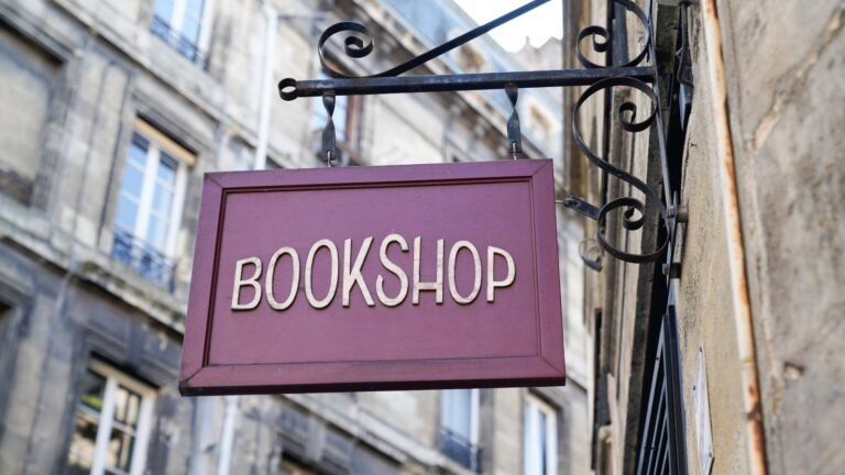 Purple bookshop sign hanging from a decorative wrought-iron bracket against a stone building, reading 'BOOKSHOP'
