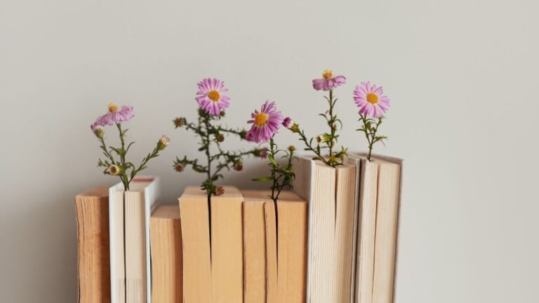 Rows of old books standing upright with pink daisy-like flowers sprouting from their pages against a neutral wall