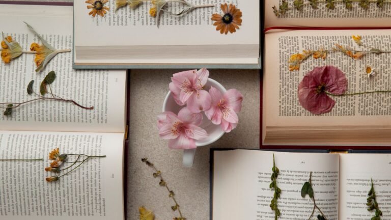 Top-down view of open books arranged around a white cup with pink flowers; dried blossoms rest on the pages.
