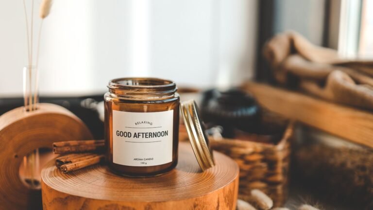Amber aroma candle labeled 'Good Afternoon' sits on a round wooden block with cinnamon sticks nearby in a cozy, spa-like setting, indoors.