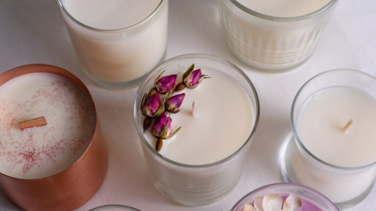 Set of white candles in glass jars on a light surface, center candle decorated with dried pink rosebuds far from the copper-toned tin filled with tiny pink speckles nearby