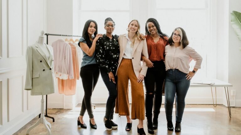 Five diverse women posing together in a bright fashion studio with a clothing rack and a mannequin in the background.