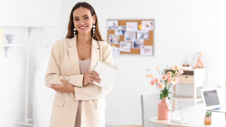 Woman in a beige blazer smiling at the camera, holding a spiral notebook in a bright office setting.