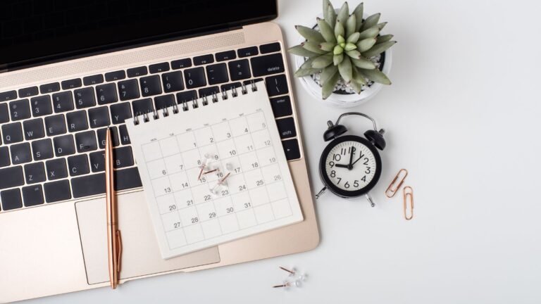 Rose-gold laptop with a desk calendar on the keyboard, copper pen, succulent plant, and a black alarm clock on a white desk.