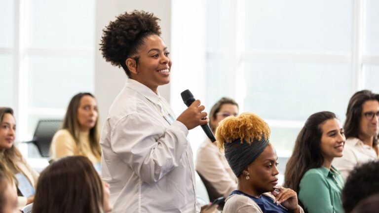 Woman with natural hair holding a microphone, speaking to a seated audience in a bright room.