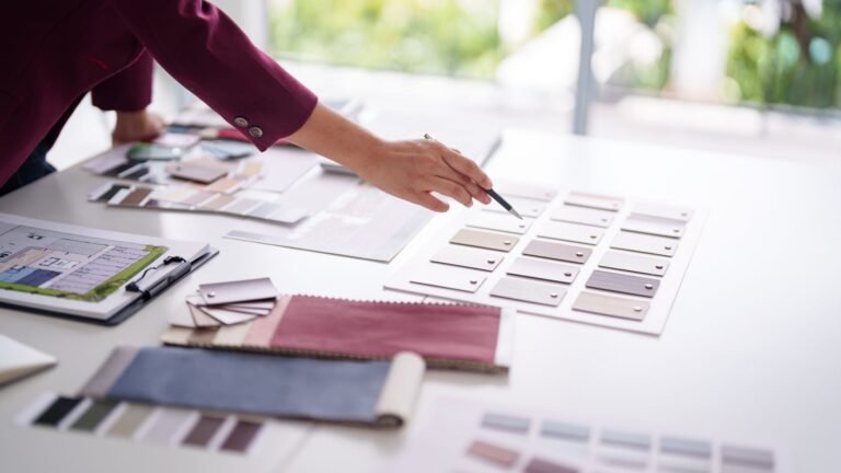 Designer’s hand selecting color and fabric swatches laid out on a white table in a bright studio.