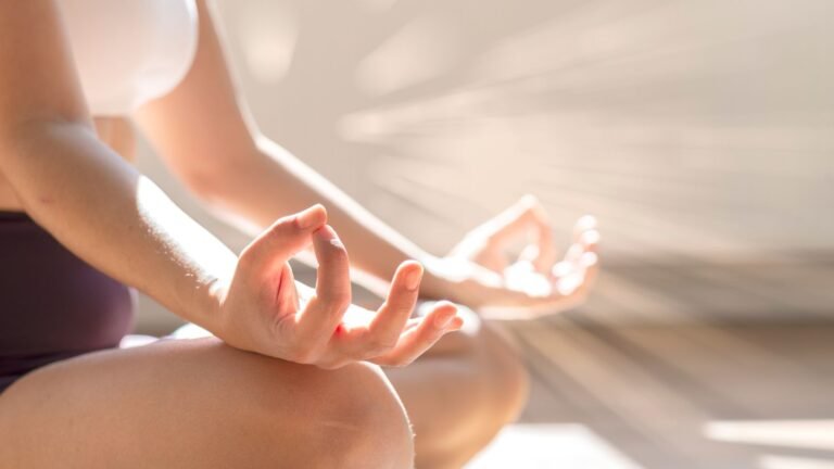 Person seated cross-legged in a meditative mudra, hands resting on knees in bright sunlight.